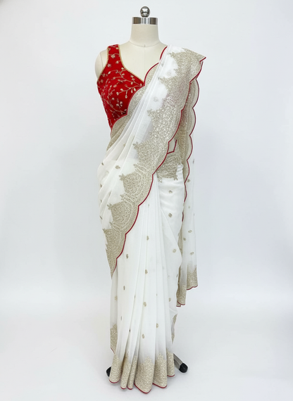 A mannequin draped in a white saree adorned with delicate golden embroidery and a scalloped glitter border outlined in red. The saree is paired with a sleeveless red velvet blouse featuring intricate threadwork, creating a refined and festive ensemble.